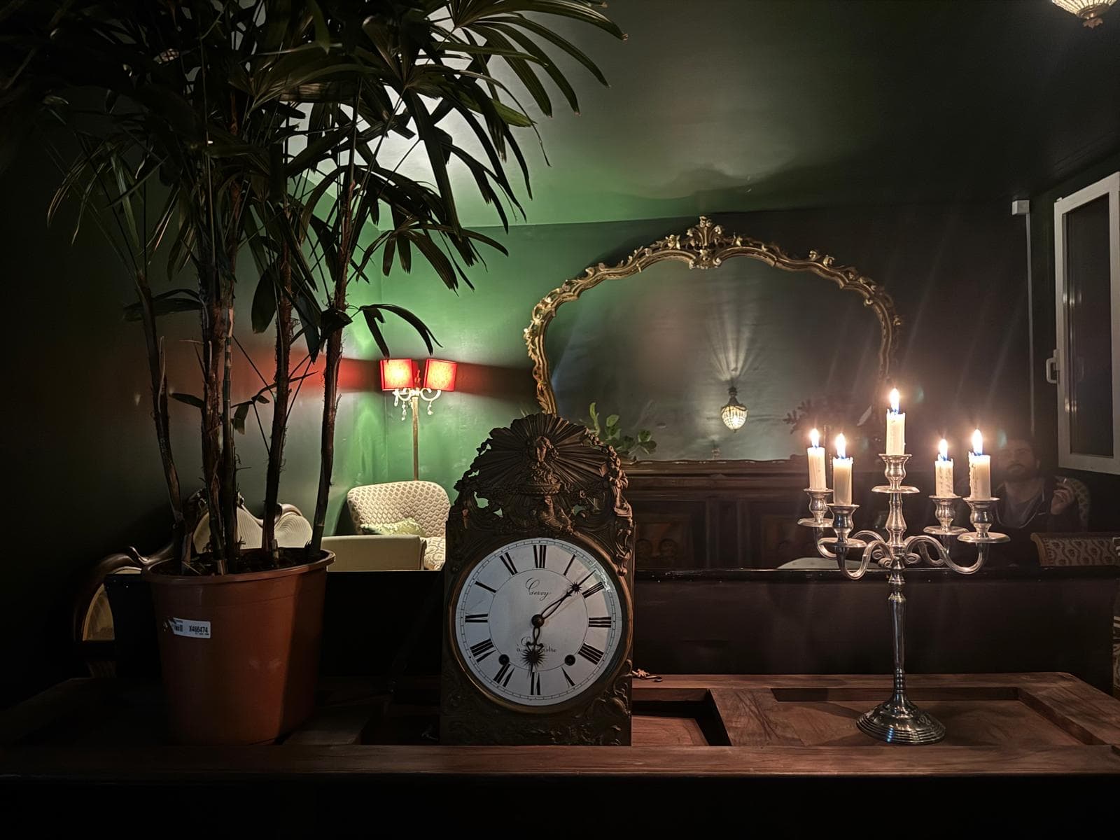 Ornate clock, lit candelabra, and large palm plant in a dimly lit green room.
