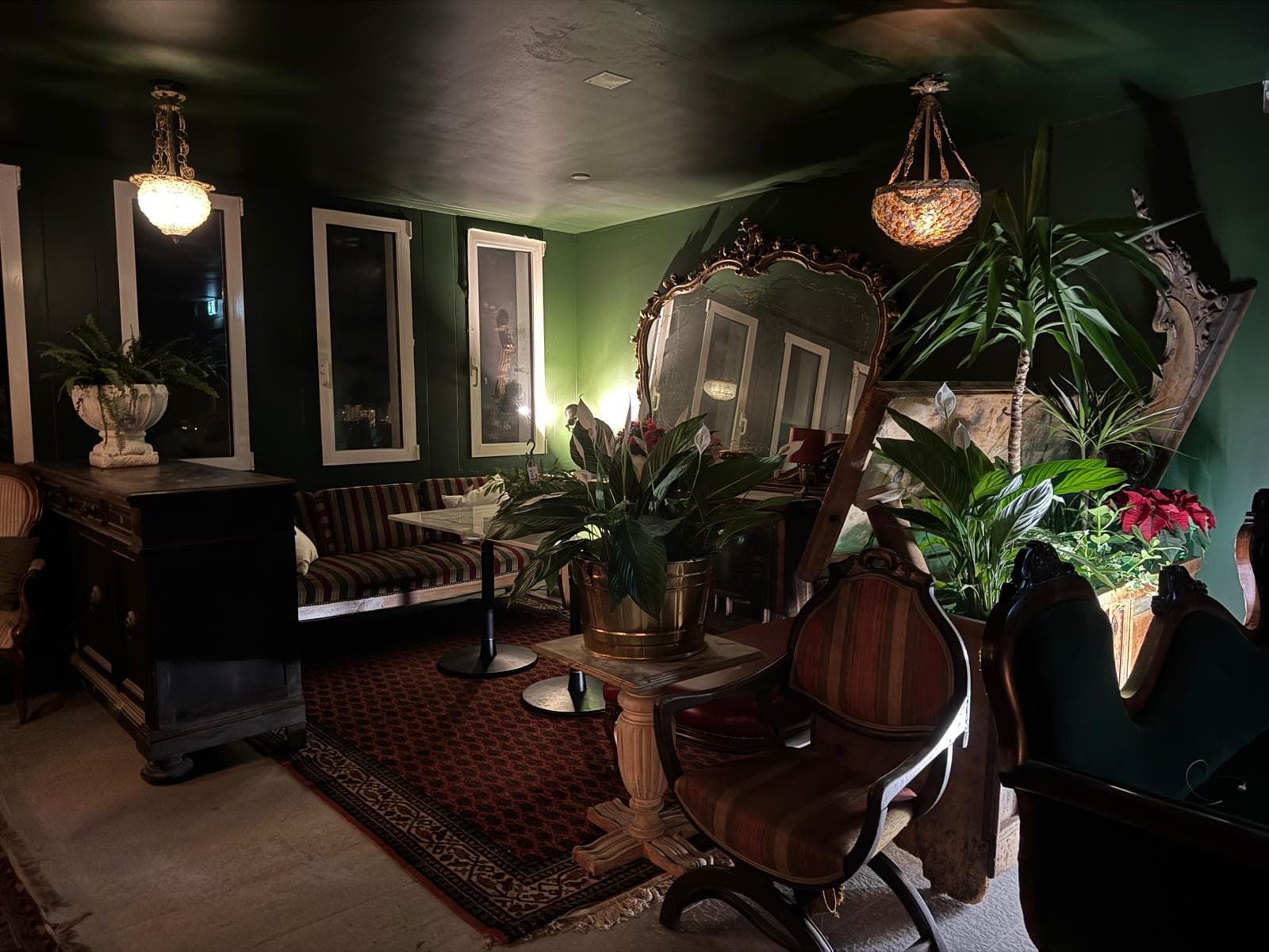 Dimly lit room with green walls, antique furniture, ornate mirror, and lush potted plants.