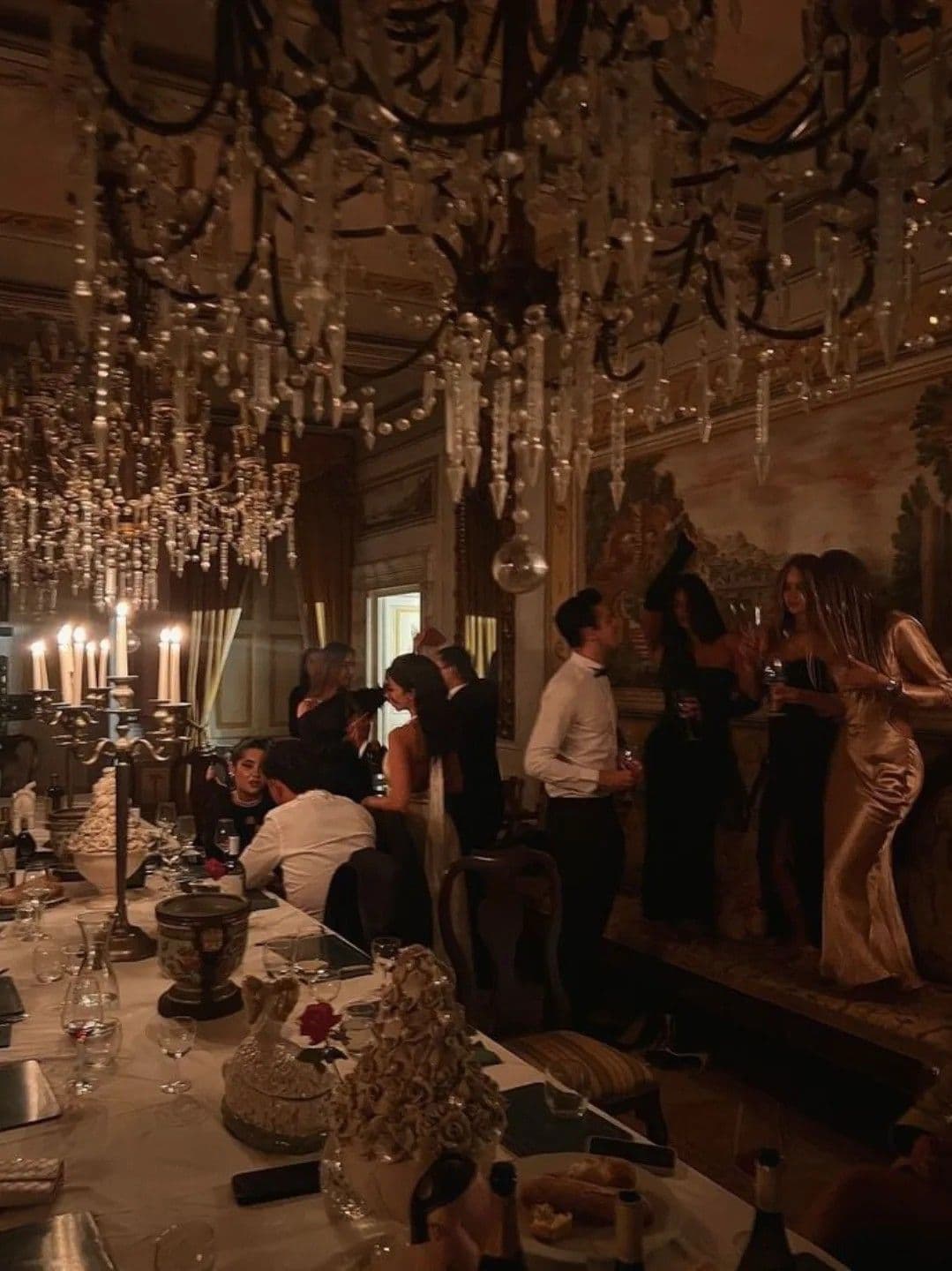 Elegantly dressed guests socialize in a grand dining room illuminated by massive crystal chandeliers.