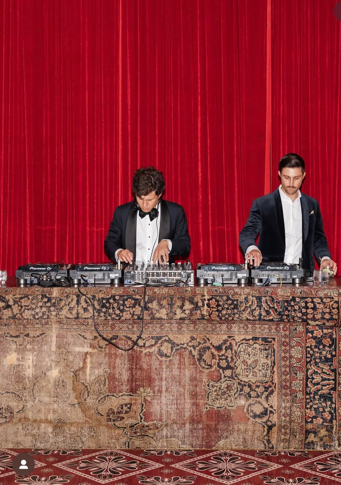 Two men in formal suits DJing behind a rug-covered table against red velvet curtains.