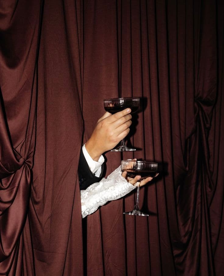 Formal hands holding coupe glasses with dark liquid emerge from behind heavy brown curtains.