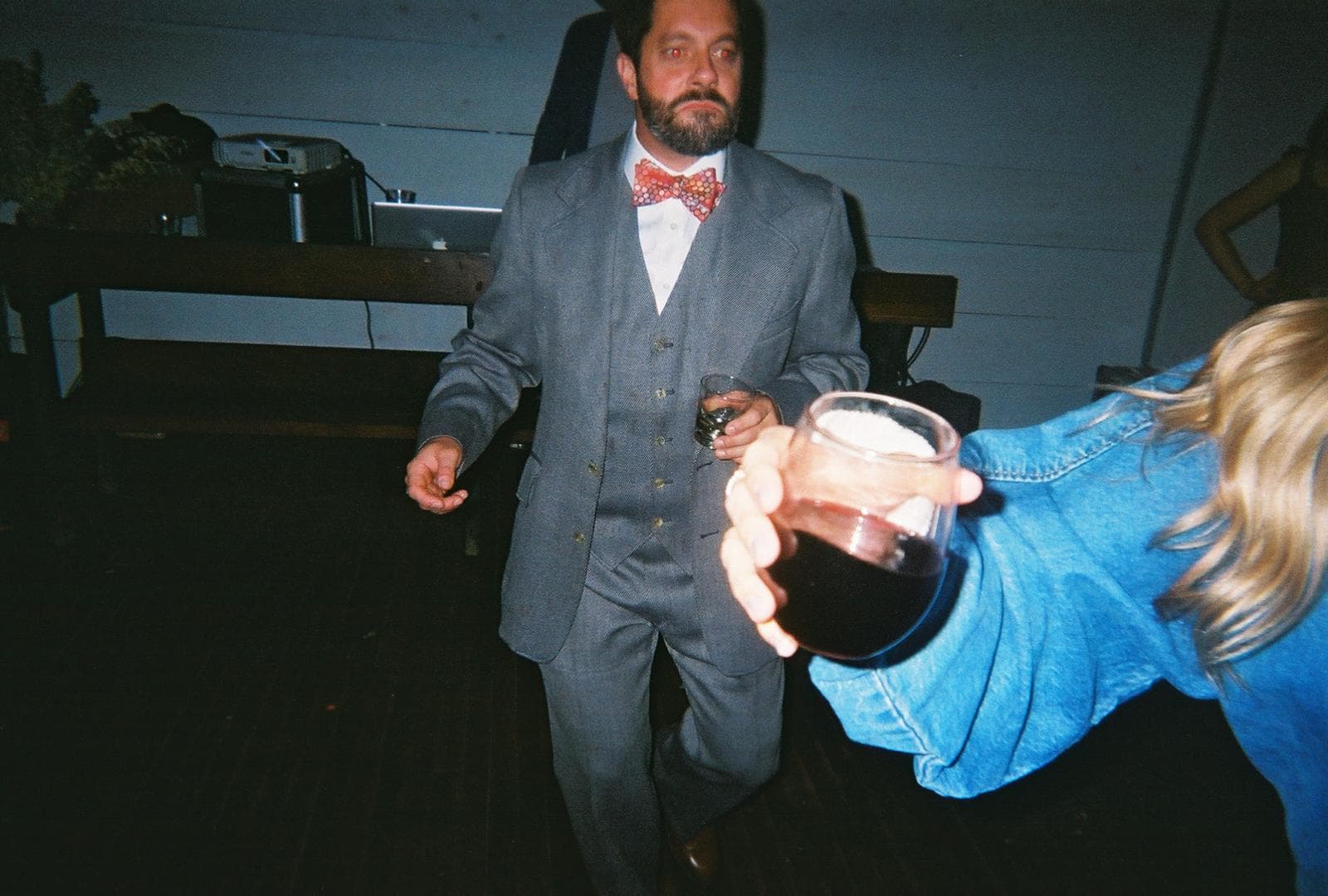 Bearded man in a grey suit and bow tie holds a drink at a party.