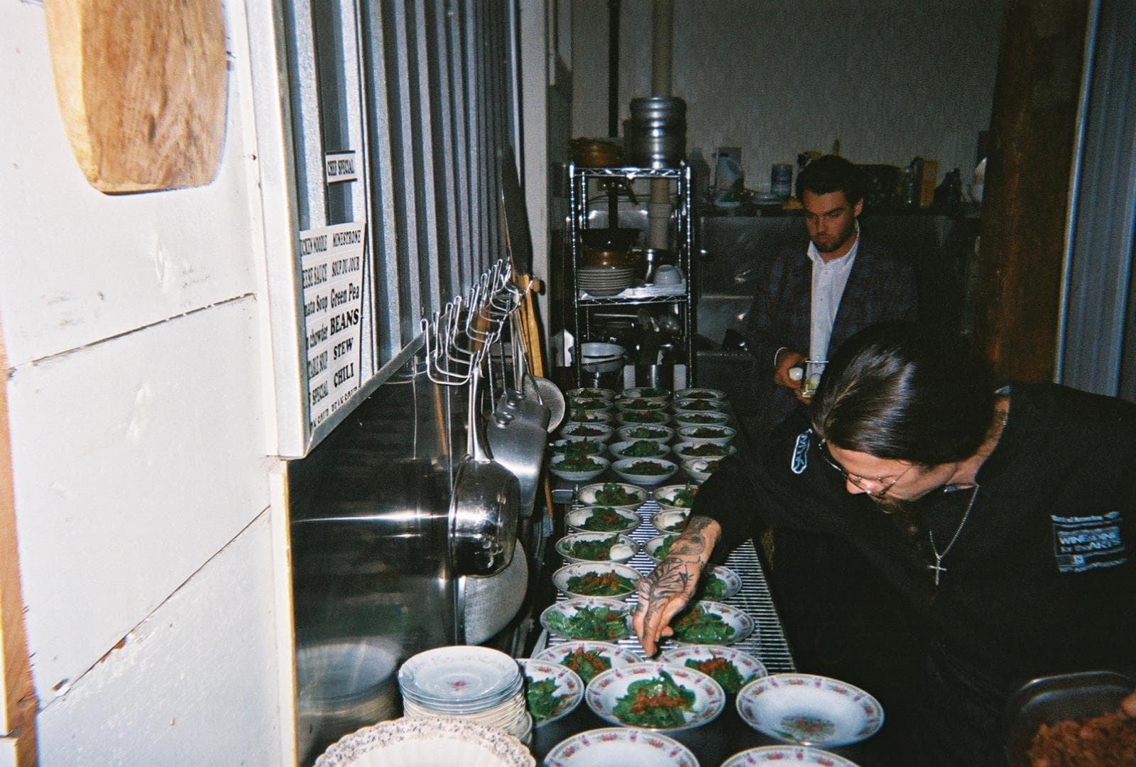 Tattooed chef garnishing rows of small salad bowls in a kitchen with hanging steel pots.