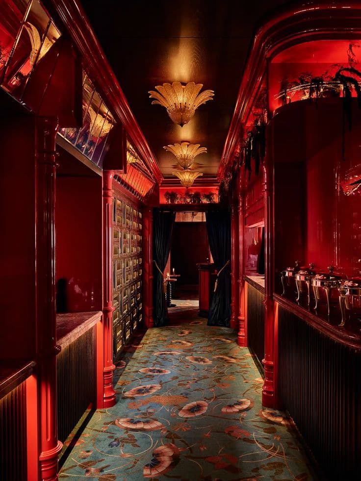 Opulent red corridor featuring glossy columns, warm floral ceiling lights, and a patterned teal carpet.