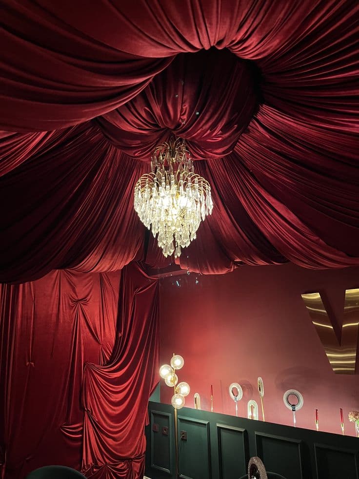 Ornate crystal chandelier hanging from a ceiling draped in luxurious, pleated red velvet fabric.