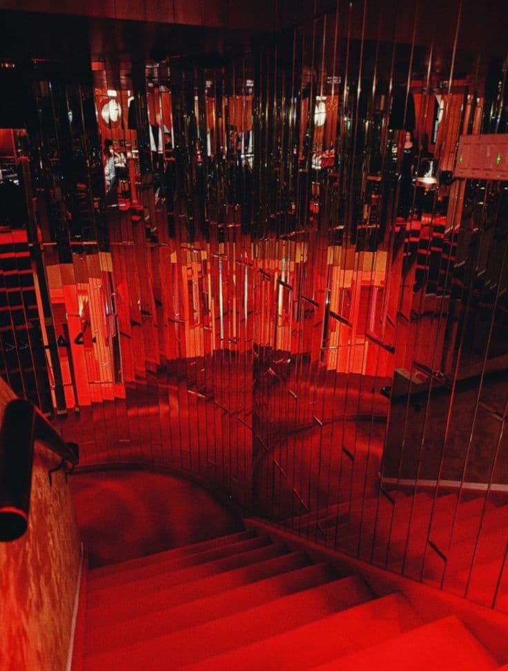Red-lit staircase descending past vertical mirrored panels creating fragmented reflections in a moody interior.